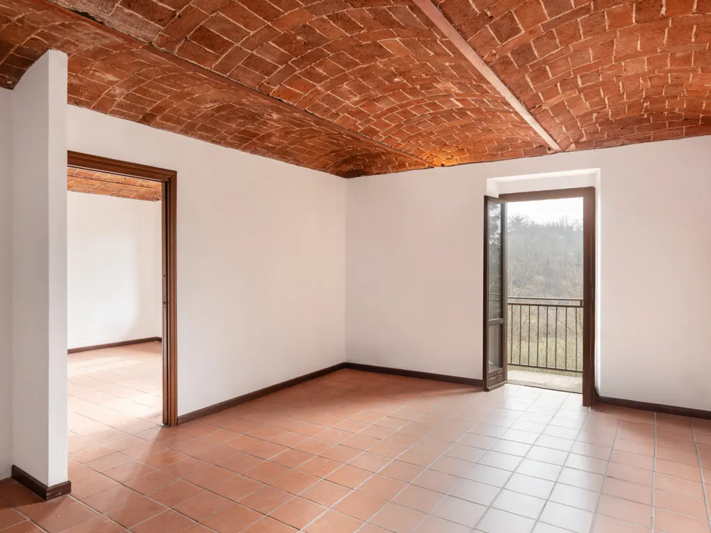 Empty room with terracotta tile floor and brick vaulted ceiling. Open door to balcony with trees in background.