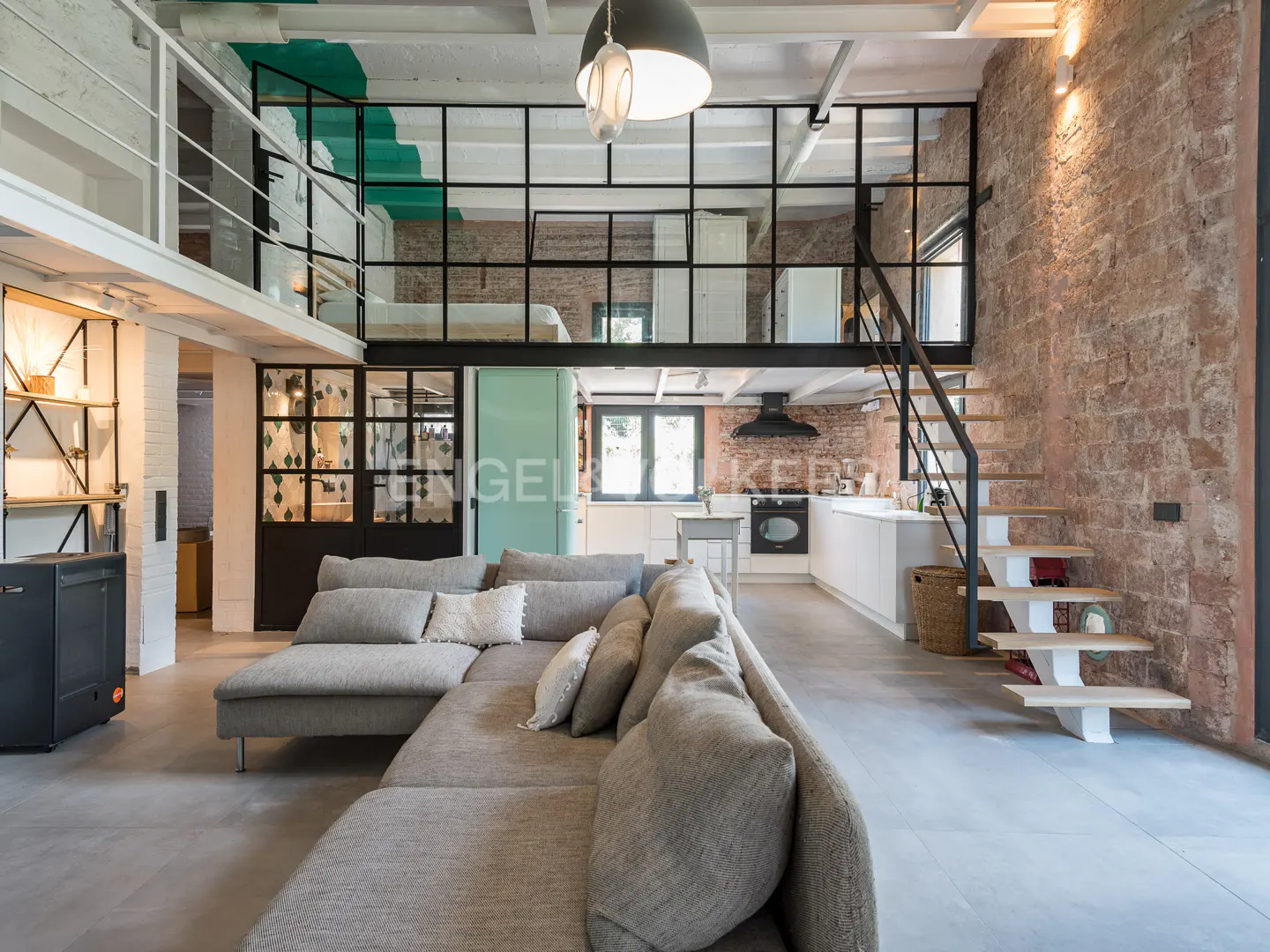 Open-concept loft with gray sectional sofa, brick wall, and black-framed mezzanine with stairs to kitchen.
