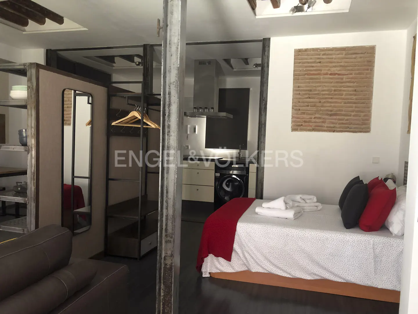 Studio apartment with a bed, kitchen, and closet. The bed has red pillows and a red blanket. The kitchen has a stainless steel hood.