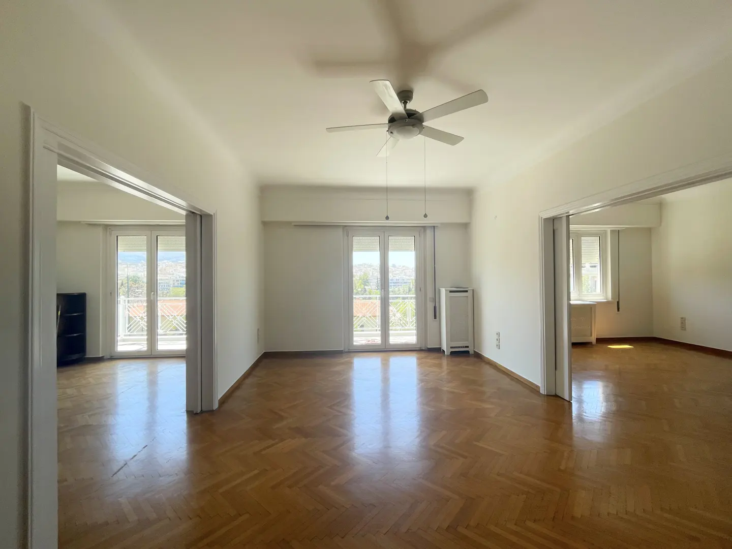 Bright, empty living room with herringbone wood floors, white walls, ceiling fan, and balcony access through glass doors.