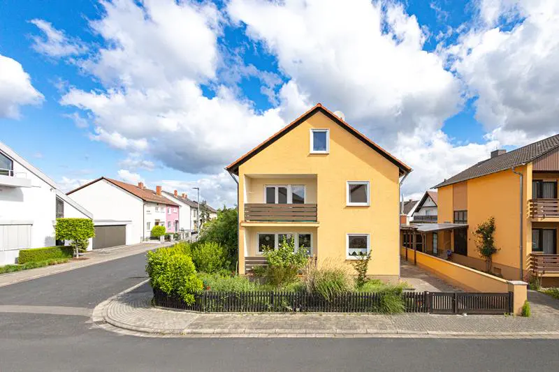 Two-story yellow house with a brown balcony, a black fence, and a blue sky with white clouds.