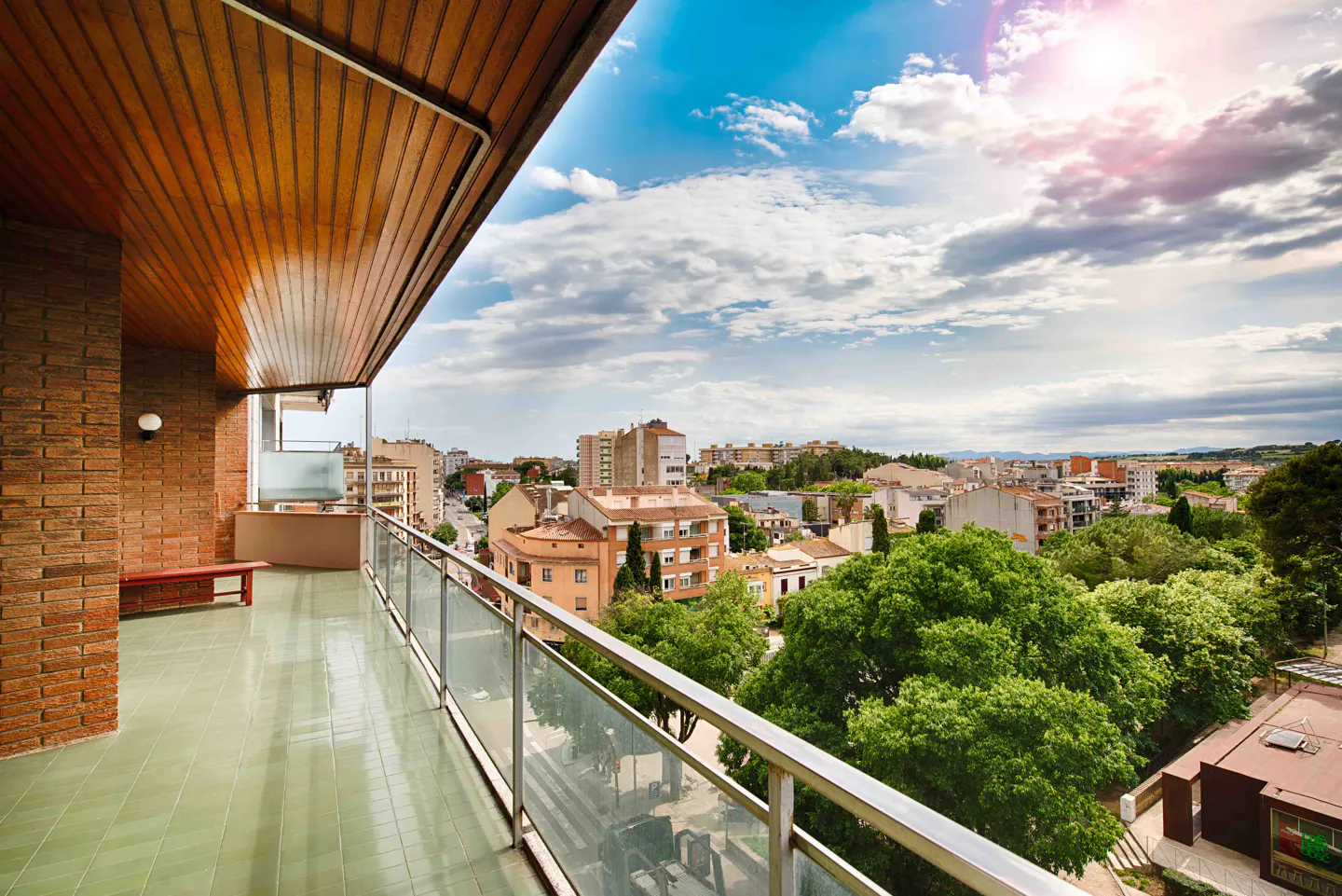 Wide balcony view with green tile floor, brick wall, wood ceiling, and glass railing overlooking a city with trees under a cloudy sky.