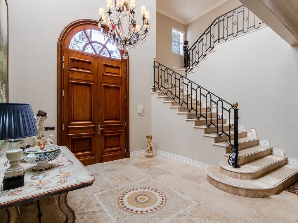Grand foyer with a wood door, crystal chandelier, and staircase with black iron railings. Beige tile flooring with a circular mosaic design.