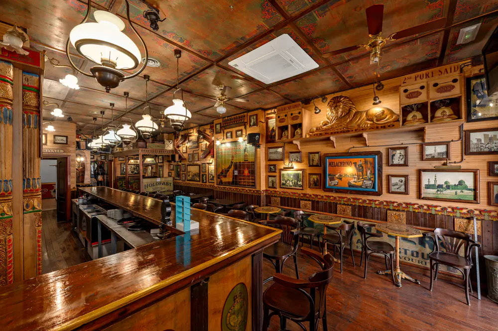 Vintage-style pub interior with a long wooden bar, hanging pendant lights, and walls decorated with framed art and a lion.