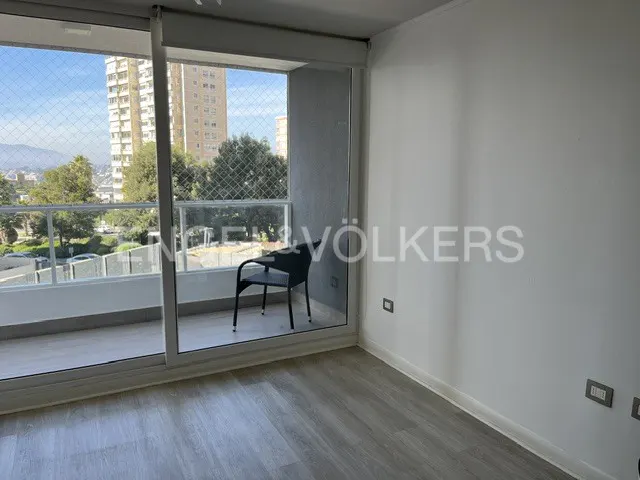 Bright room with gray wood floors, white walls, and a balcony with a black chair overlooking city buildings and trees.
