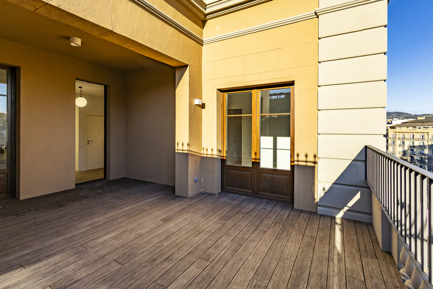 A sunlit balcony with wooden floors, a metal railing, and a view of city buildings in the distance.