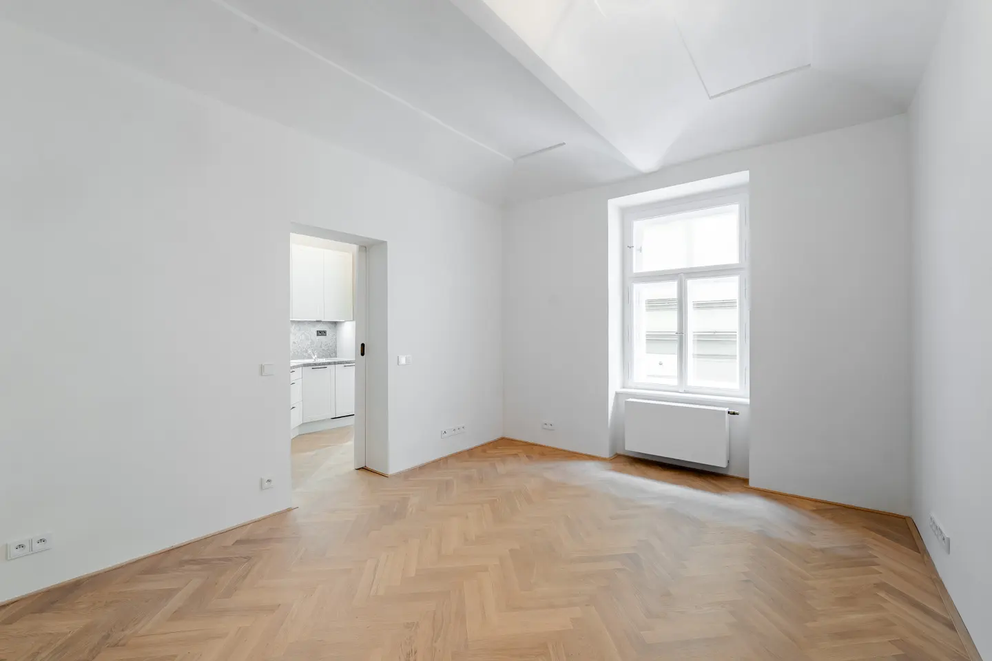 Bright, empty room with white walls, herringbone wood floor, and a window. A doorway leads to a white kitchen.