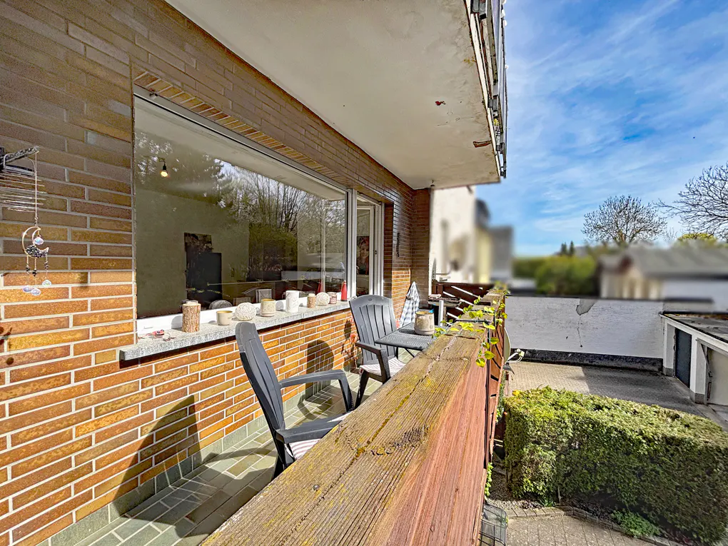 Balcony view with two black chairs and a small table. A brick wall and a large window are behind the chairs. The sky is blue with clouds.