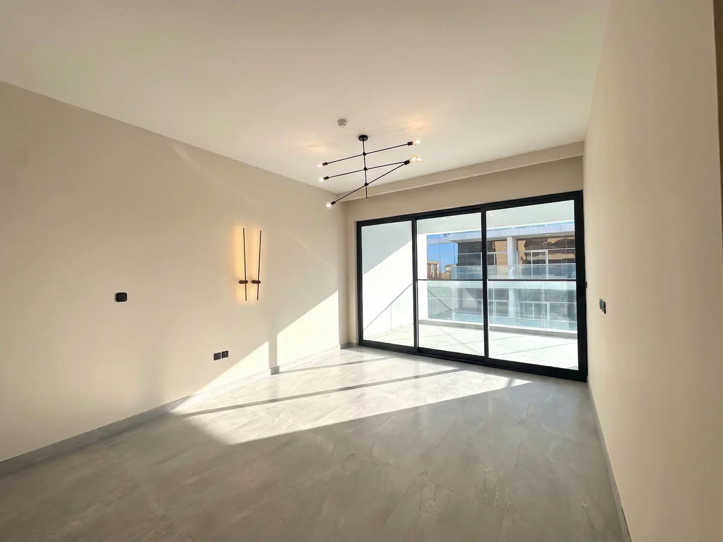 Empty room with beige walls, modern chandelier, wall sconces, and large sliding glass doors leading to a balcony.