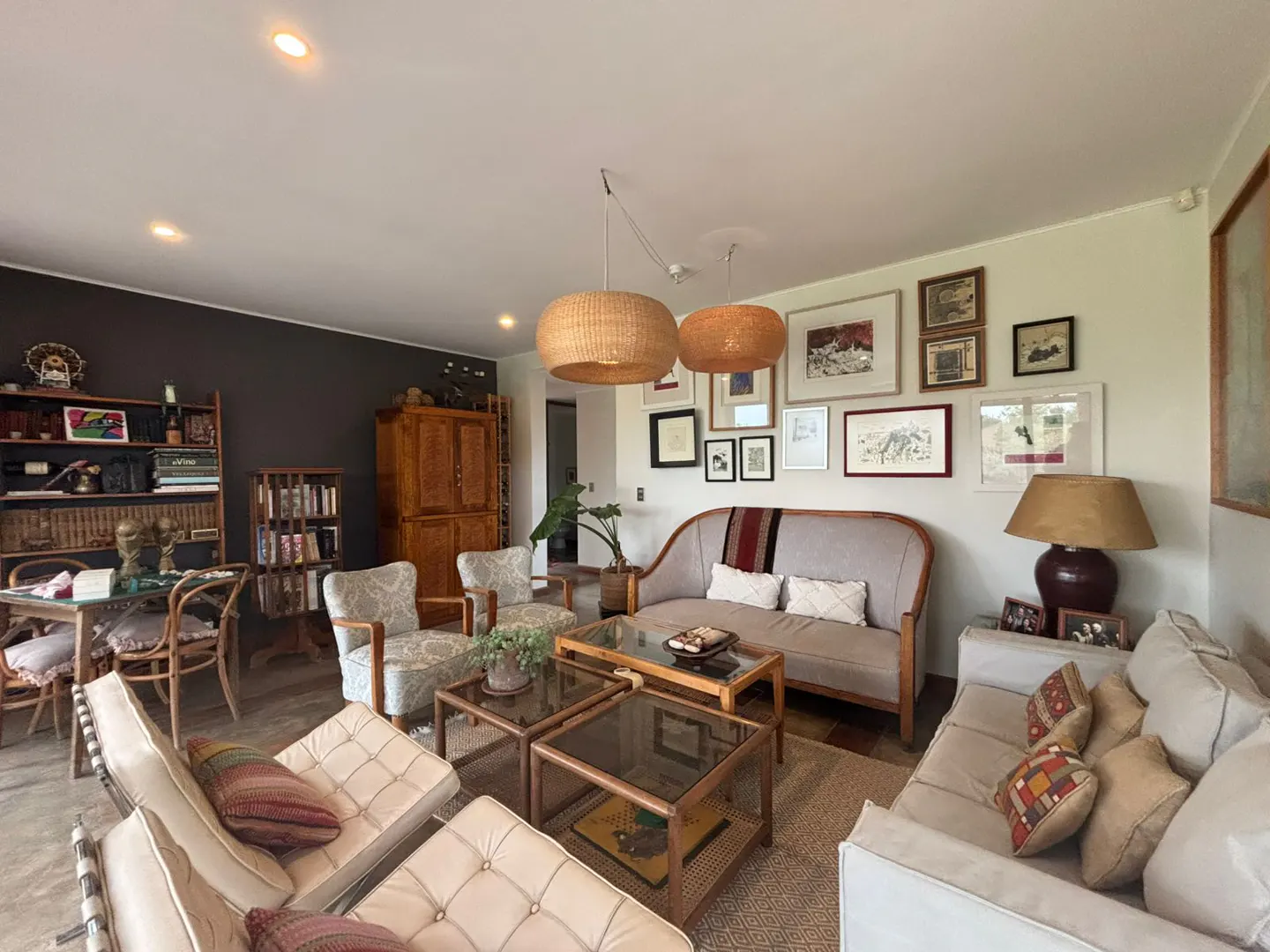 Living room with sofas, chairs, and tables. Wicker pendant lights hang above. A gallery wall displays framed art. Bookshelves line the back wall.