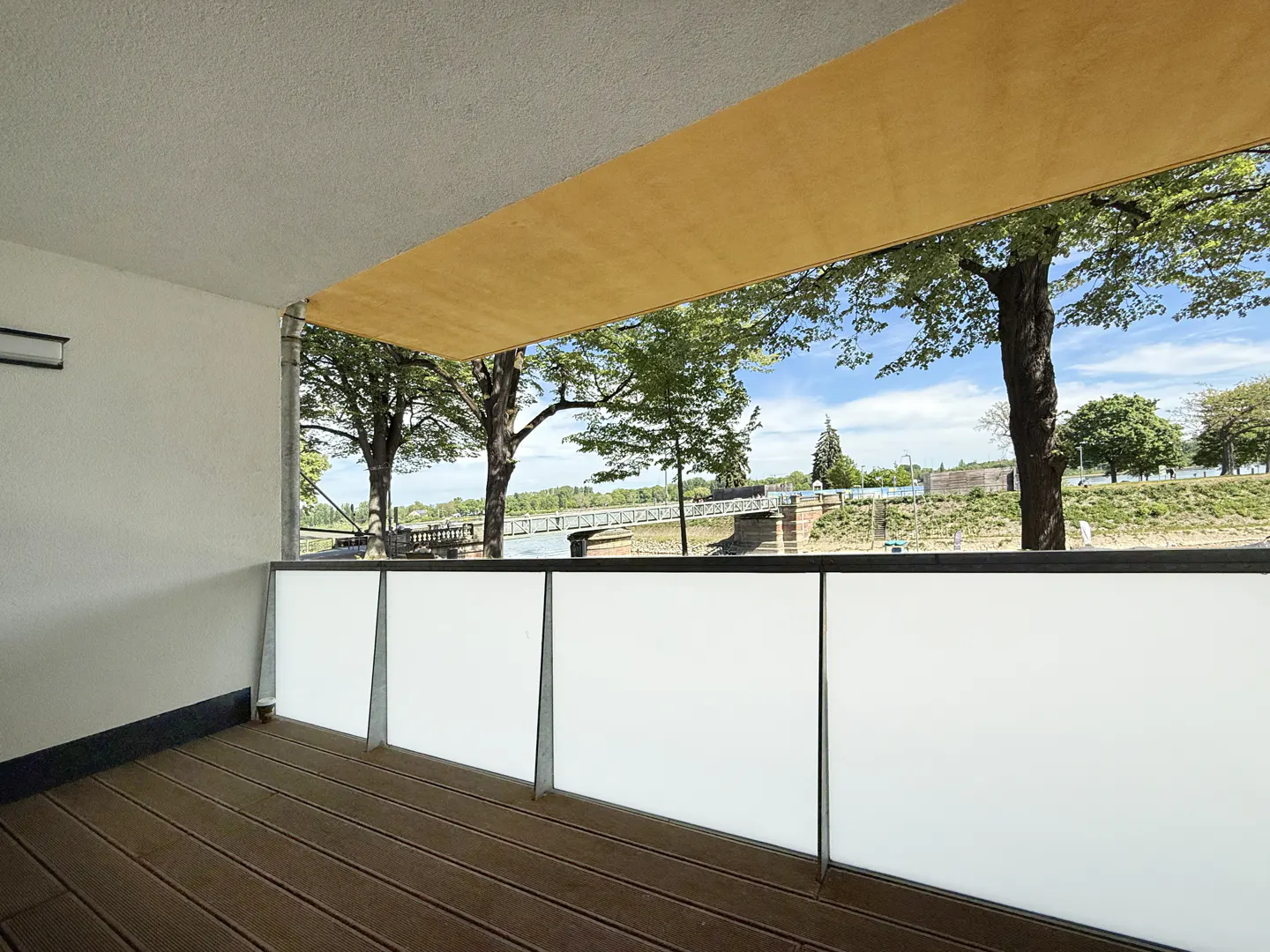 Balcony view with brown wood floor, white panels, and a bridge over water in the background. Trees and blue sky are visible.