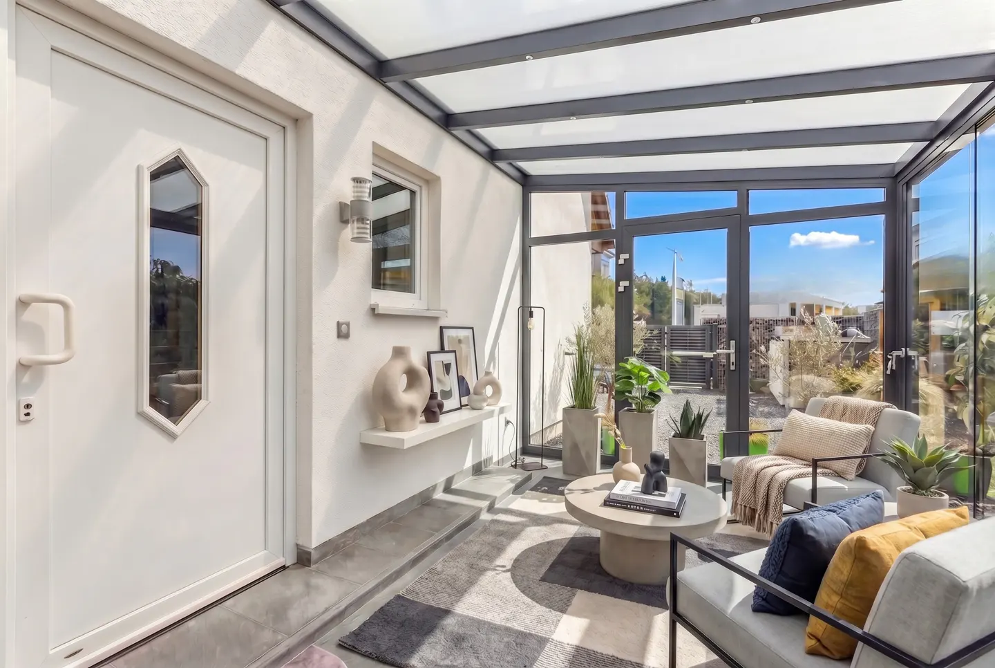 Bright sunroom with glass ceiling and walls, featuring two gray chairs, a round coffee table, and potted plants. A white door is on the left.