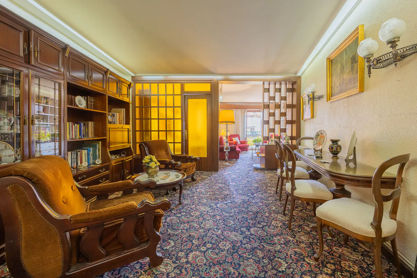 A vintage living room with brown armchairs, a bookshelf, and a floral carpet. A yellow glass door leads to another room with red furniture.