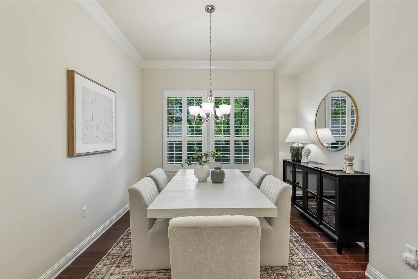 Bright dining room with a white table, upholstered chairs, and a black sideboard with lamps and a round mirror.