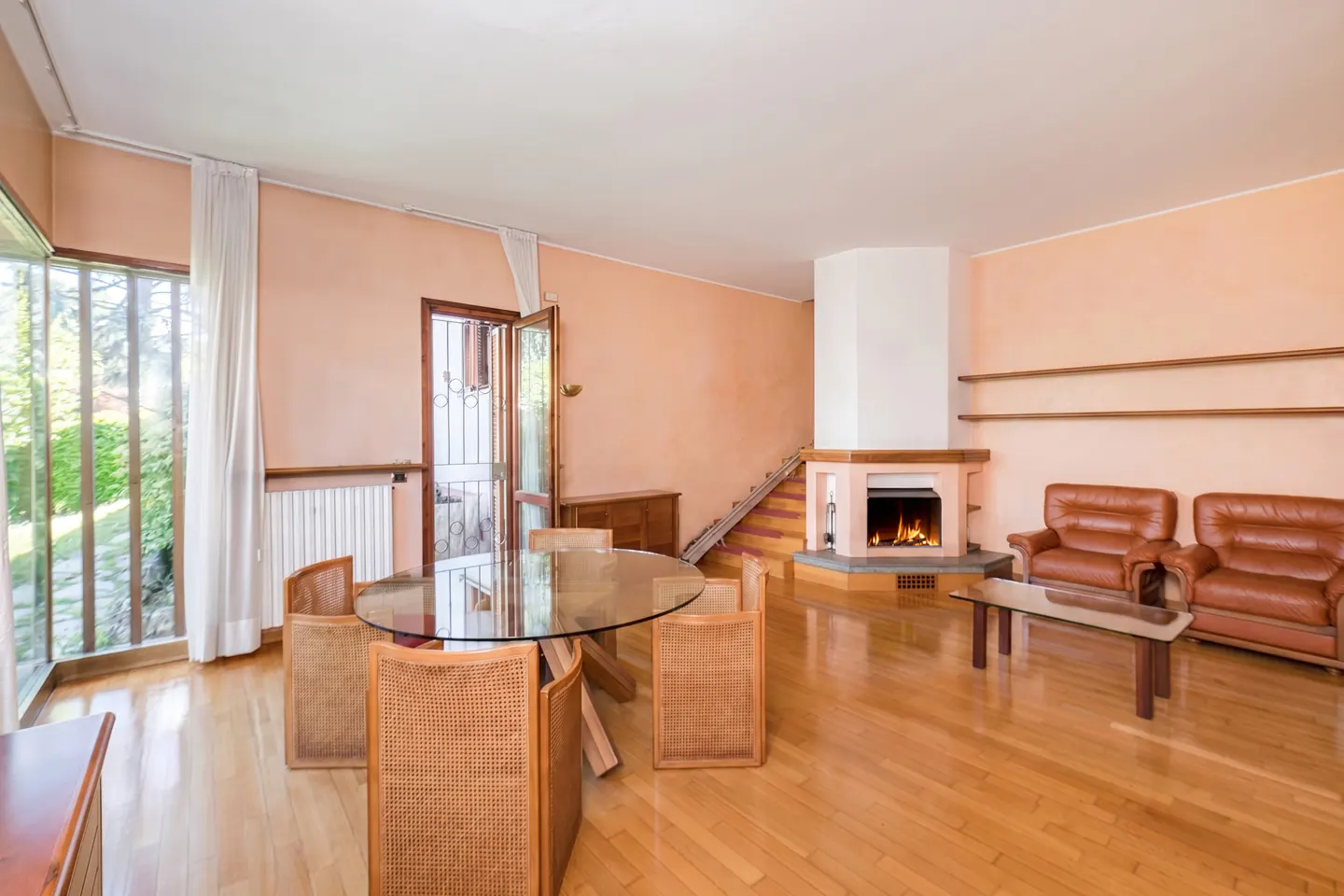 Living room with peach walls, hardwood floors, glass dining table, rattan chairs, leather sofas, and a lit fireplace.