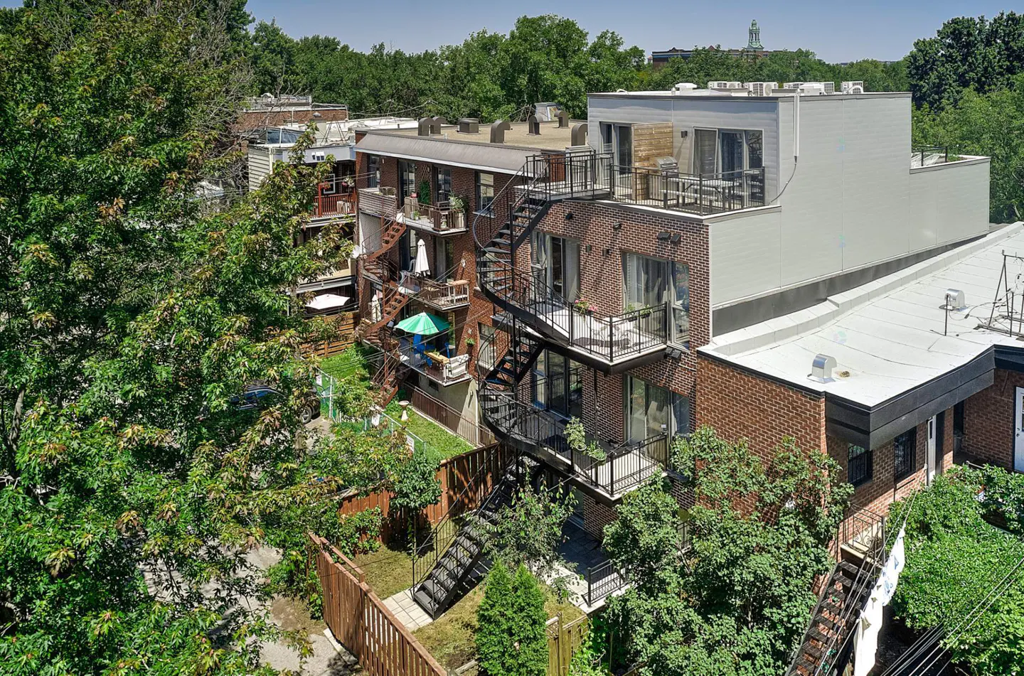 Aerial view of a brick apartment building with black metal fire escapes and balconies, surrounded by green trees.