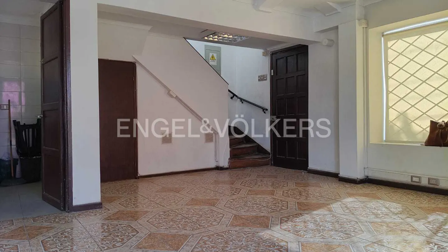 A real estate photo shows a room with brown patterned tile, white walls, a staircase, and a window with a brown bag on the sill.