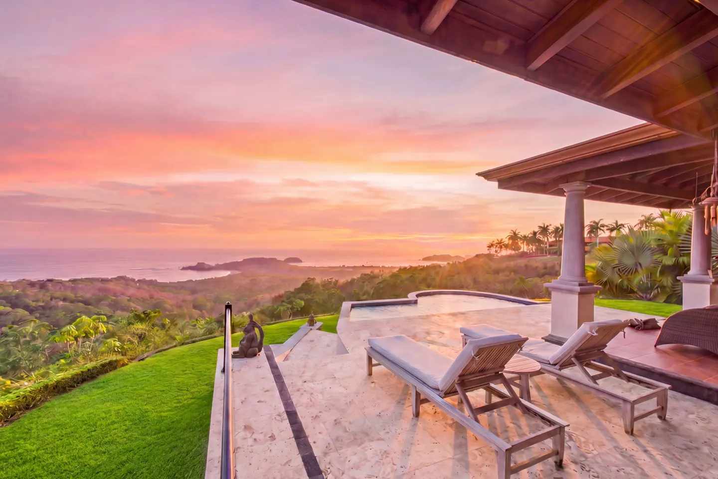 Luxury patio with lounge chairs overlooking ocean at sunset. Lush green landscape and palm trees in the background.