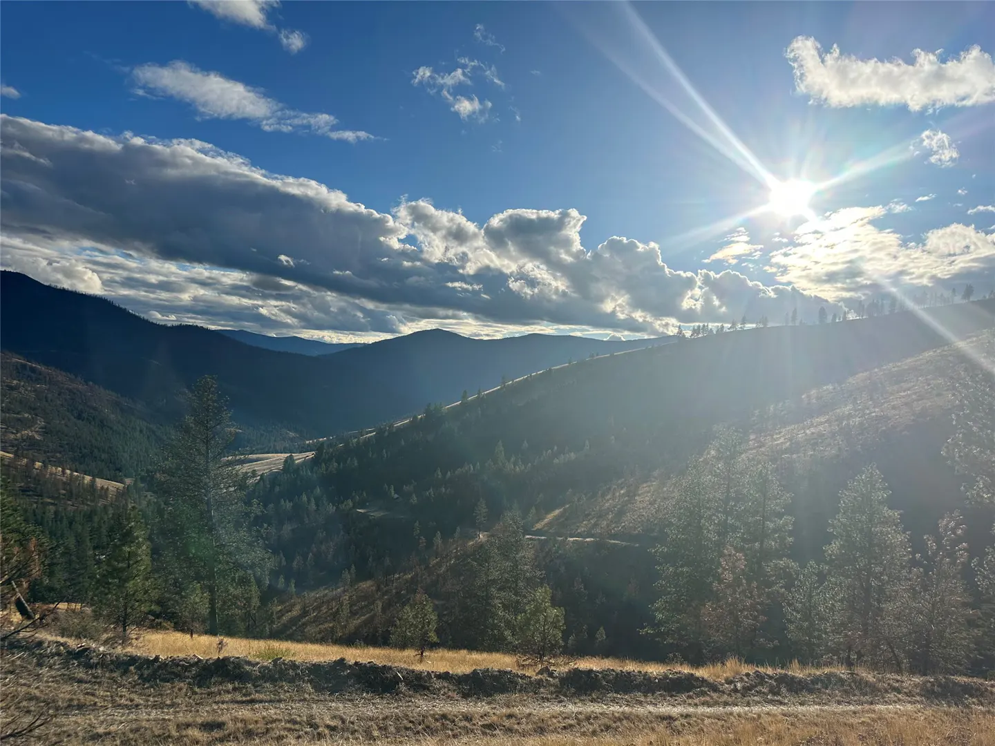 Scenic mountain view with rolling hills, scattered trees, and a bright sun shining through a partly cloudy blue sky.