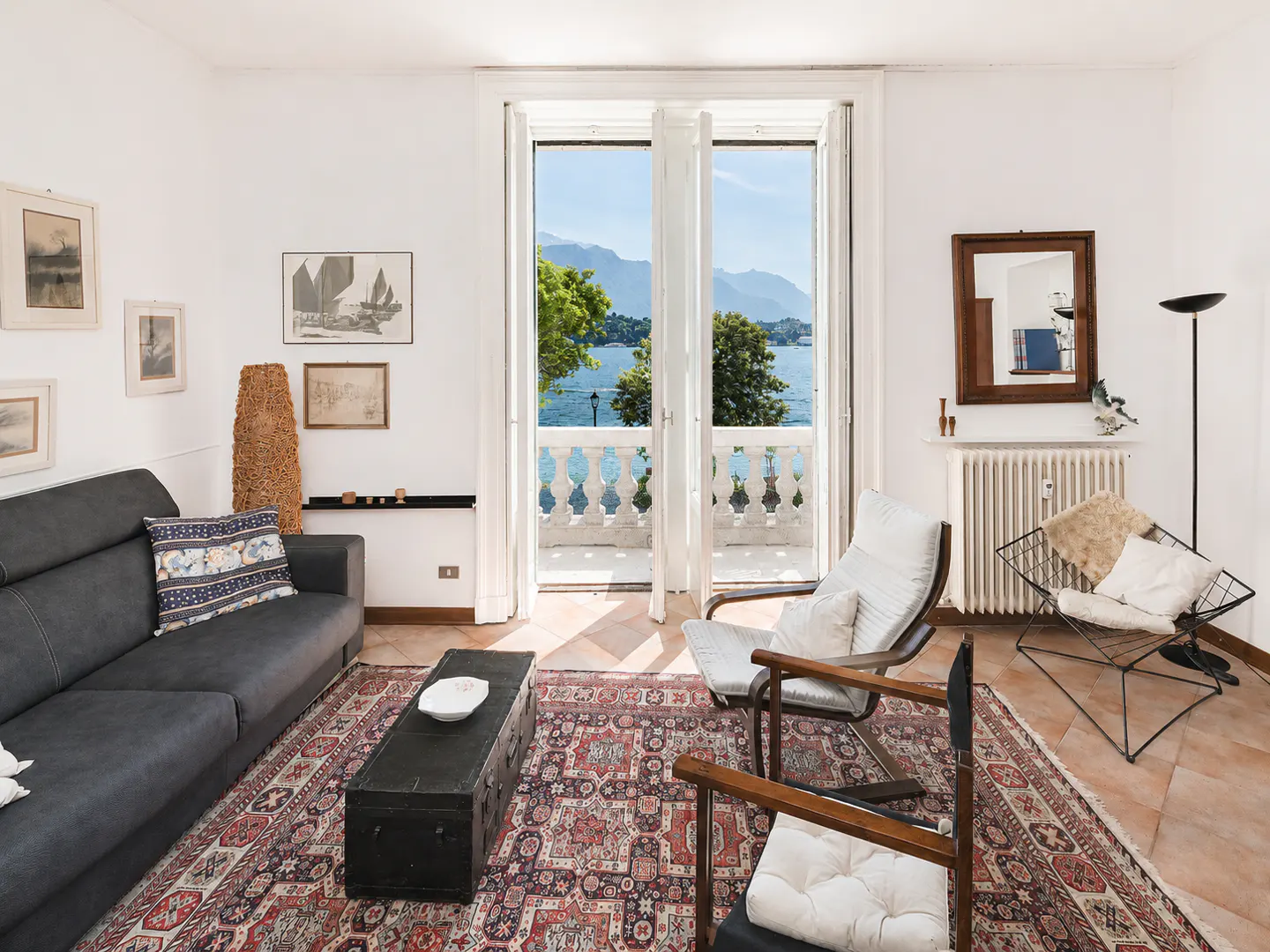 Bright living room with a gray sofa, wooden chairs, and a patterned rug. Open doors lead to a balcony with a view of a blue lake and mountains.