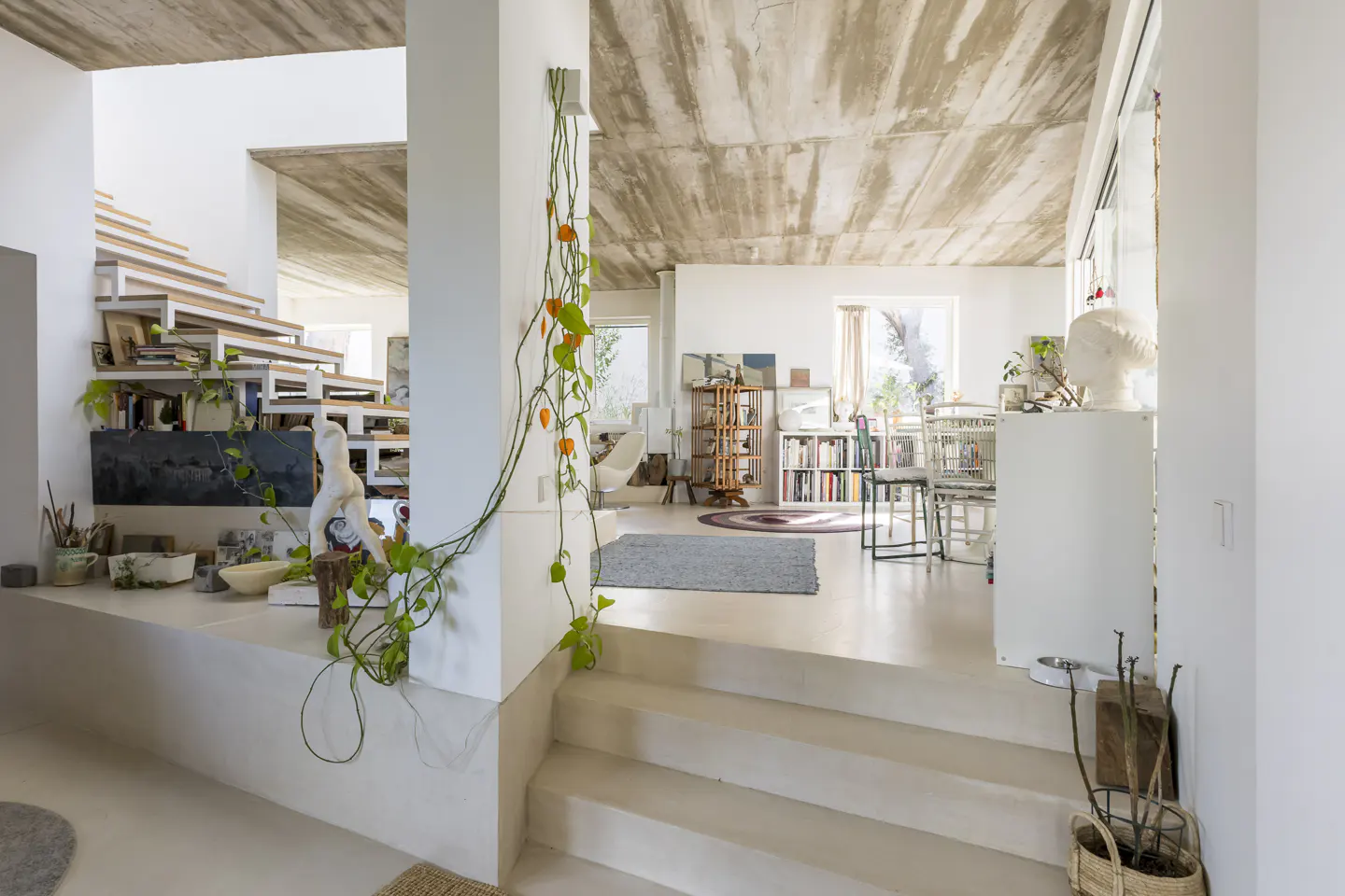 Modern living room with concrete ceiling, white walls, floating staircase, and a trailing plant on a central pillar.