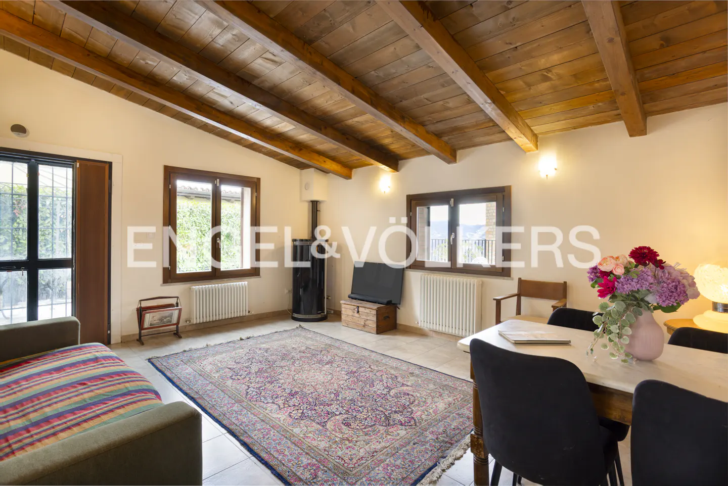 Living room with wood-beamed ceiling, sofa, dining table, and Persian rug. Windows let in natural light.