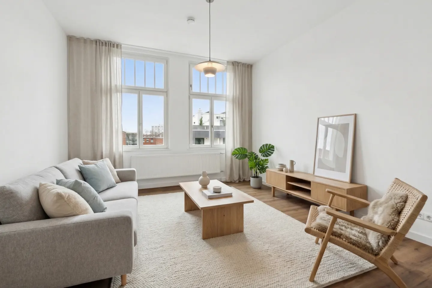 Bright living room with a gray sofa, wooden coffee table, and woven chair on a white rug. Large windows with beige curtains let in natural light.