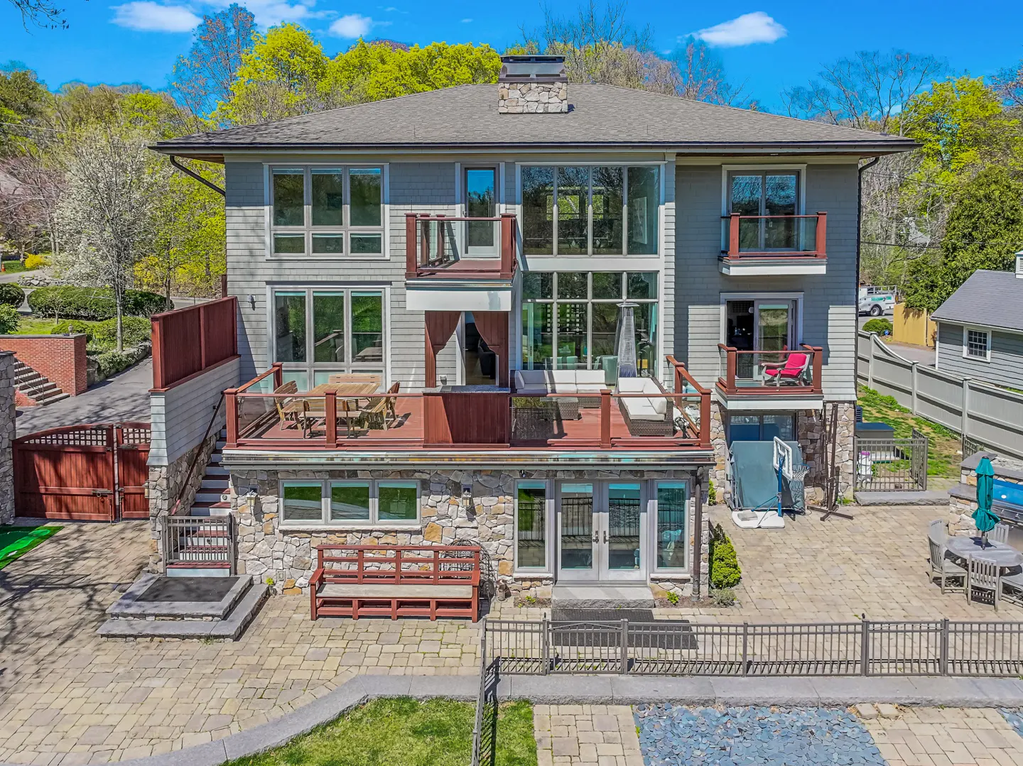 Two-story gray house with stone accents, multiple decks, and large windows. A patio with furniture and a pool are visible.