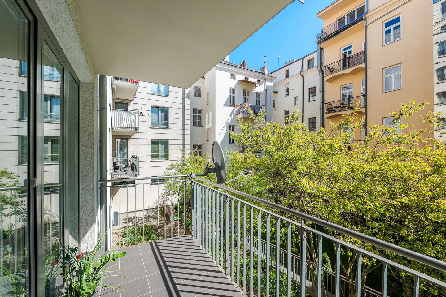 Balcony view with gray tile floor, metal railing, and satellite dish. Buildings and green trees in the background.
