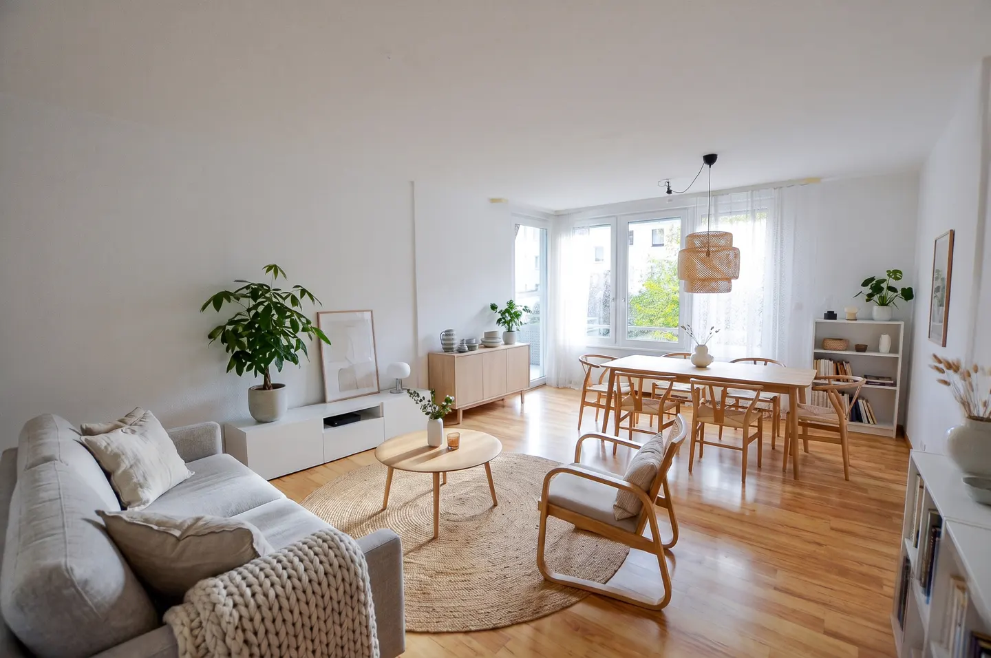 Bright, modern living room with a gray sofa, round jute rug, and wooden dining set near a window. Plants add a touch of nature.