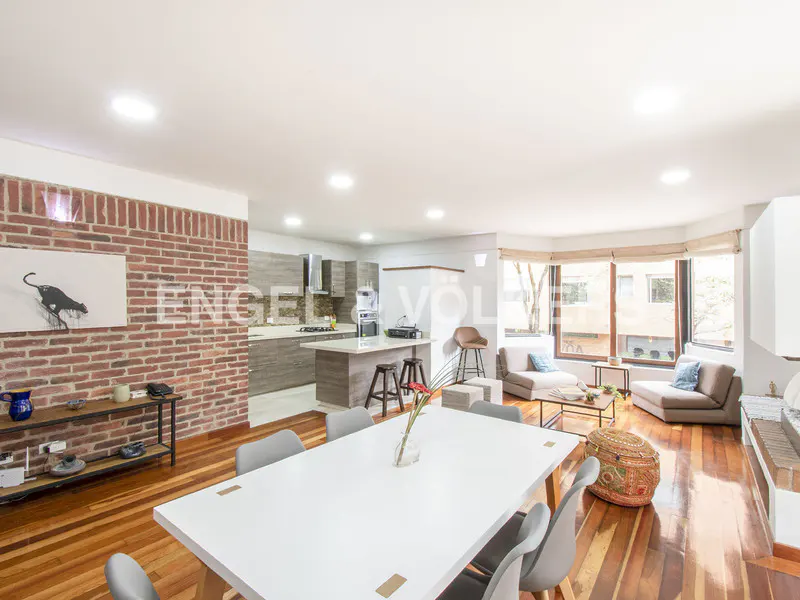 Open-concept living space with hardwood floors, brick accent wall, white dining table, and modern kitchen. Natural light fills the room.