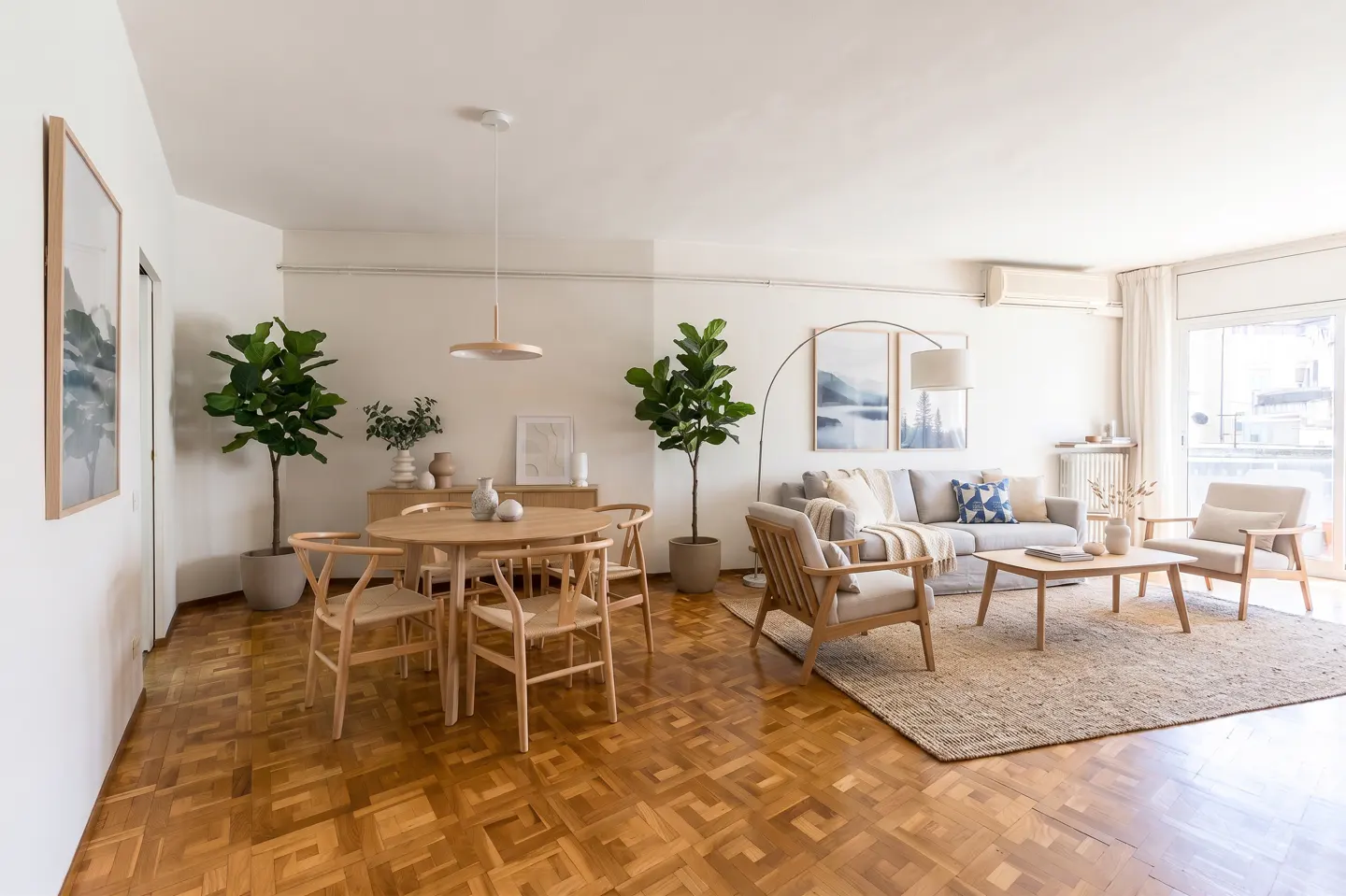 Bright, open-concept living and dining area with wood floors, light walls, and neutral furniture. Plants add a touch of green.
