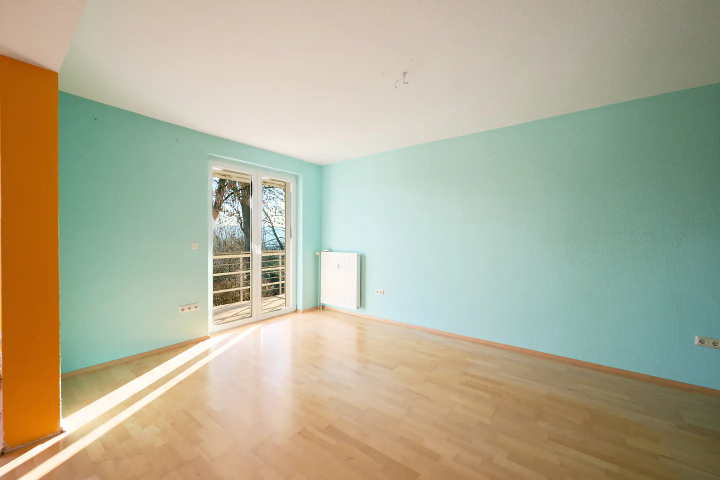 Empty room with light wood floors, light blue walls, and a white-framed glass door to a balcony. An orange column is on the left.