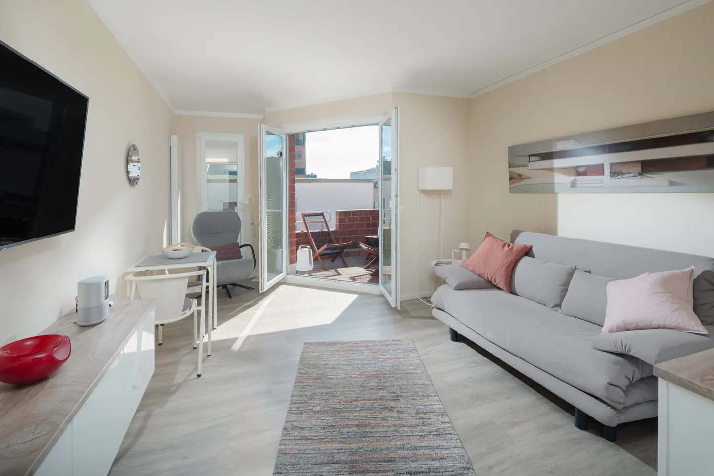 Bright living room with a gray sofa, pink pillows, and open doors to a balcony with chairs. A TV is on the left wall.
