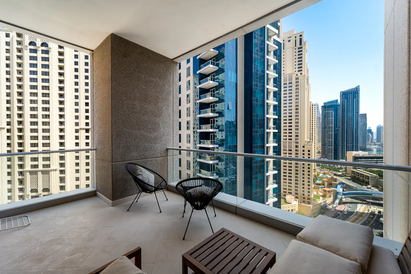 Balcony view of Dubai skyline. Two black chairs and a wooden table sit on the tiled balcony. Tall buildings are visible in the background.