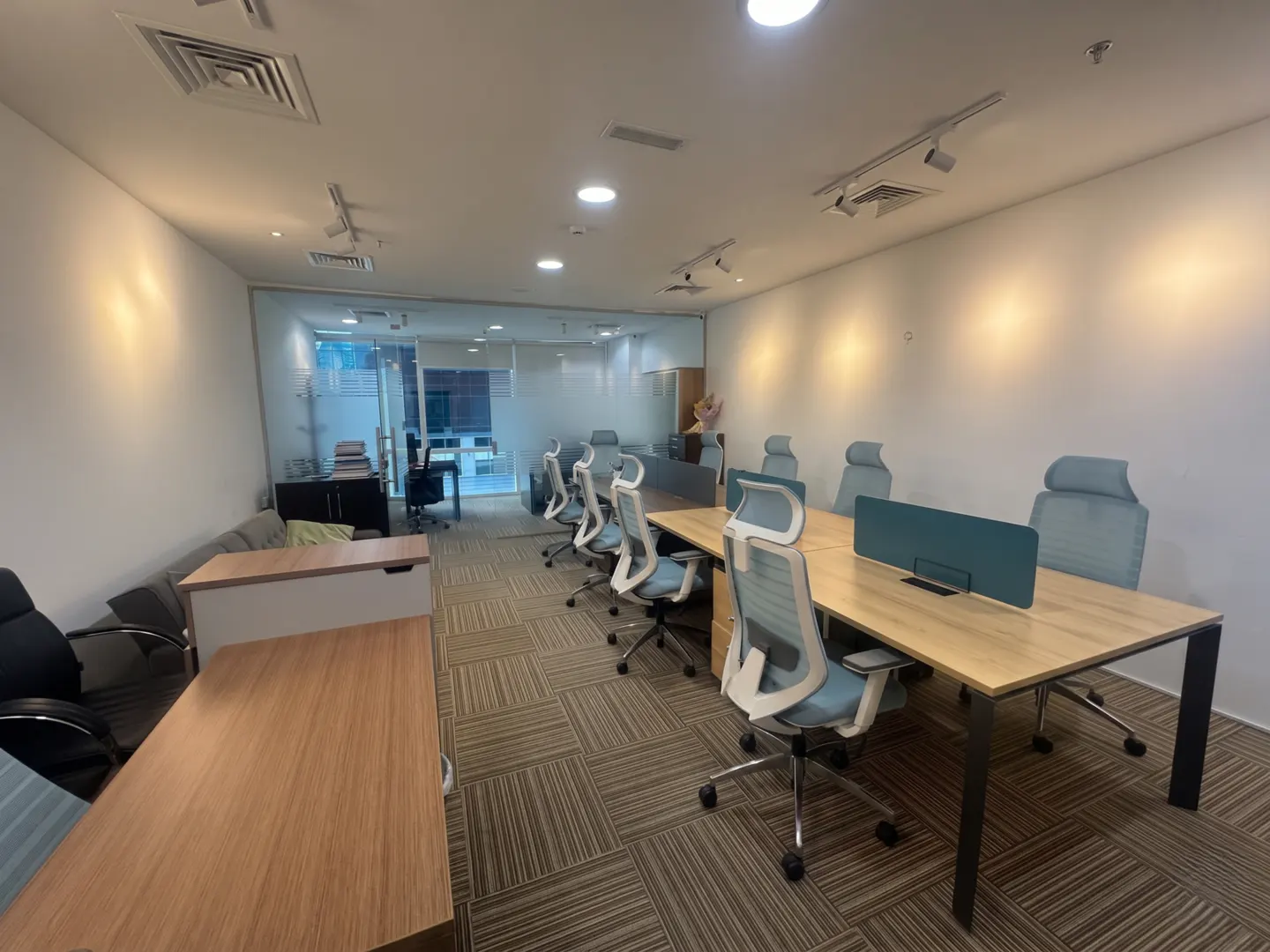 Bright office space with desks, blue chairs, and a glass-walled room in the background. The floor is carpeted in a brown geometric pattern.