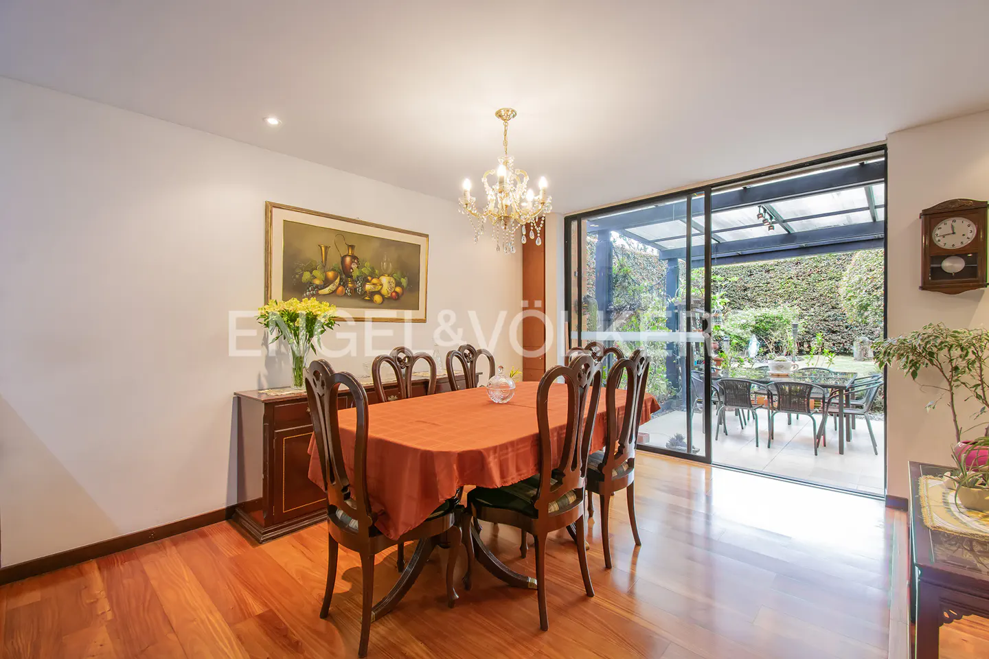 Dining room with wood floors, table with orange cloth, and chairs. Sliding glass doors lead to a patio with green foliage.