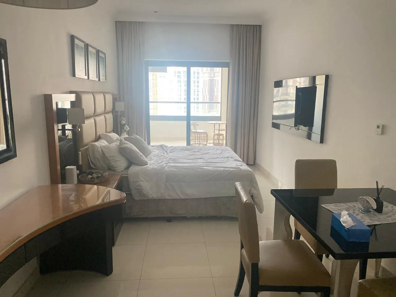 A bright studio apartment with a bed, desk, dining table, and balcony with city views.