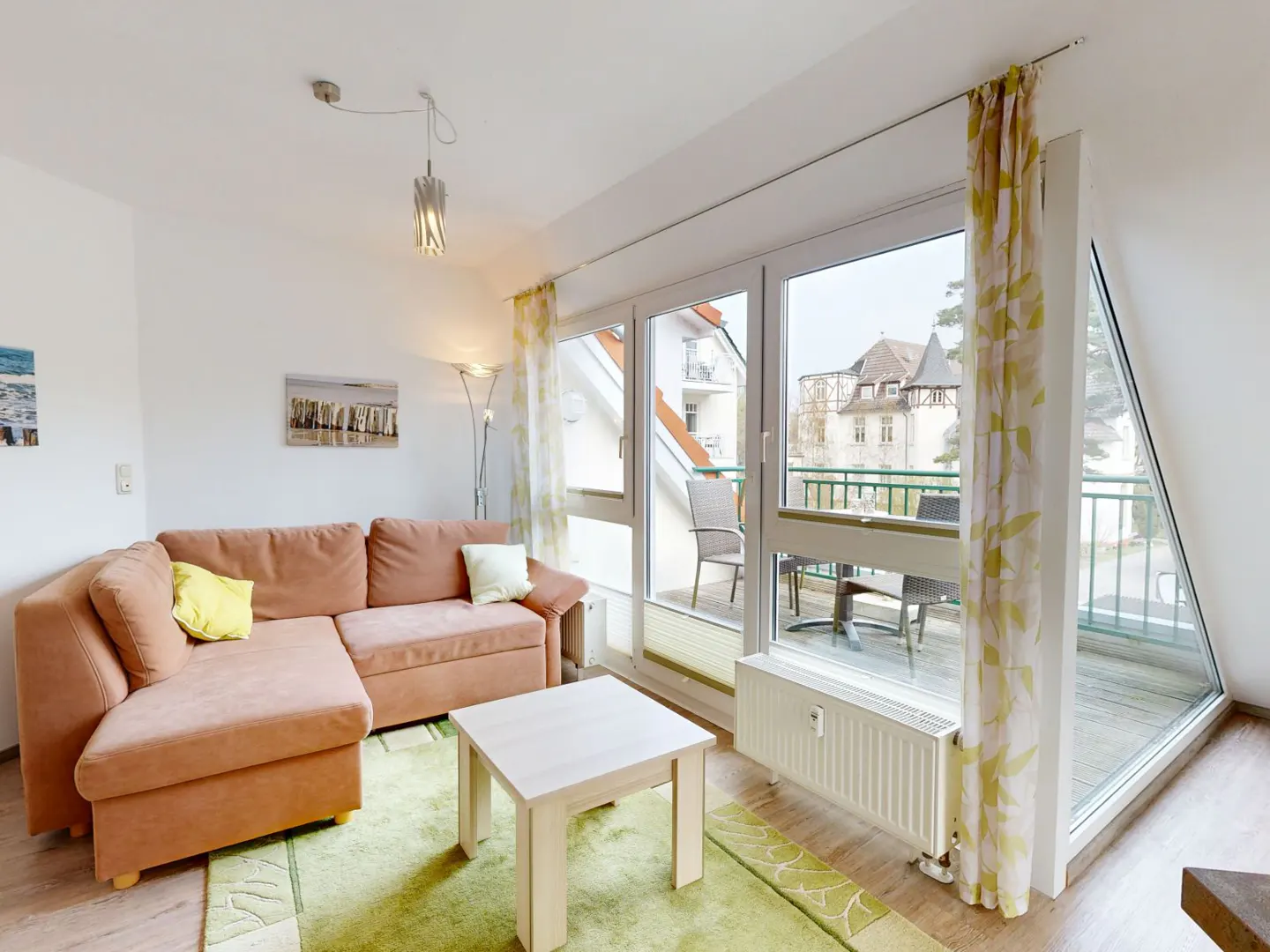 Living room with a salmon-colored sectional sofa, a white table, and a green rug. Balcony with chairs and a table is visible through the glass doors.