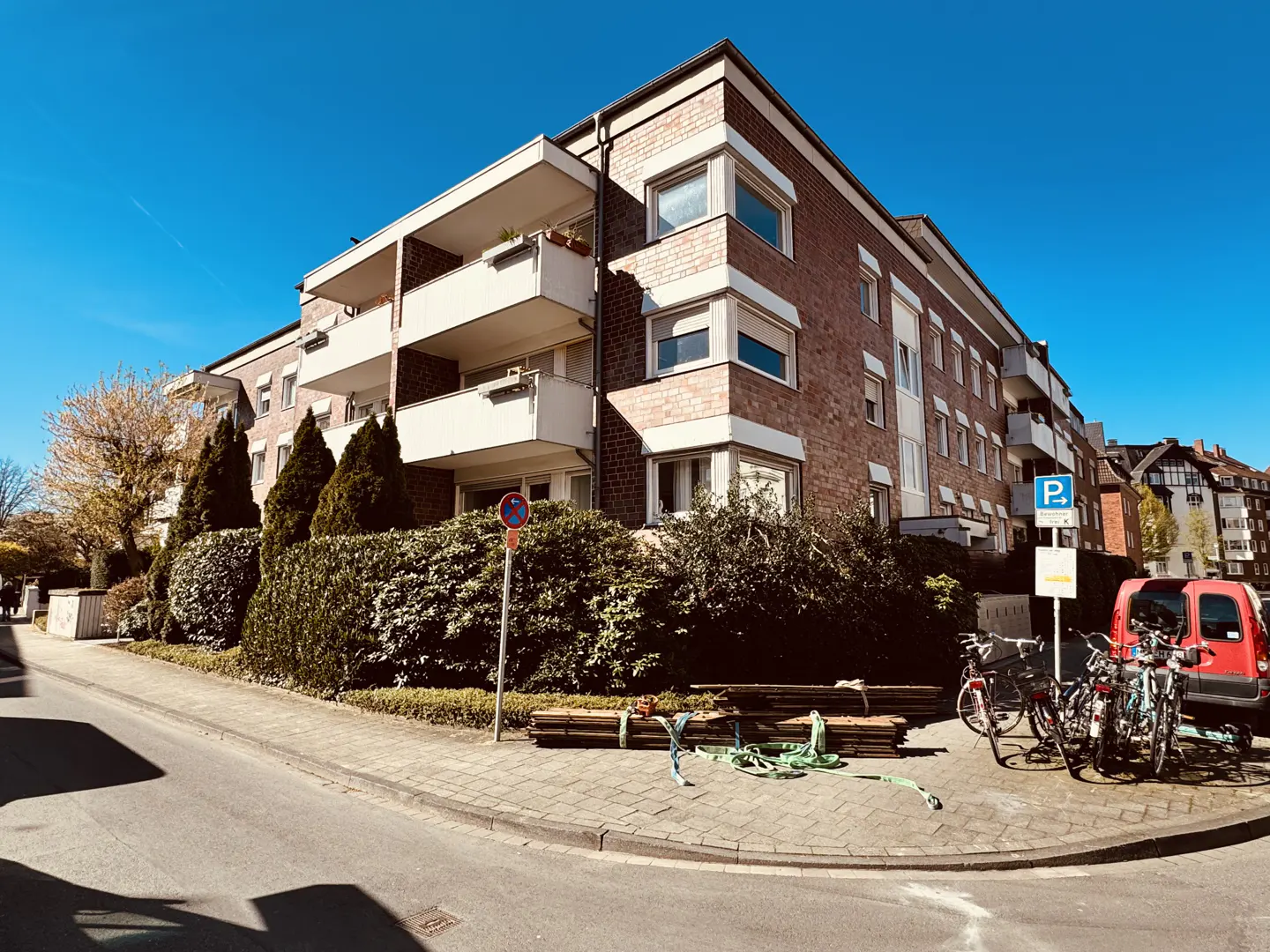 A three-story brick apartment building with white balconies under a clear blue sky. Bicycles and a red van are parked nearby.