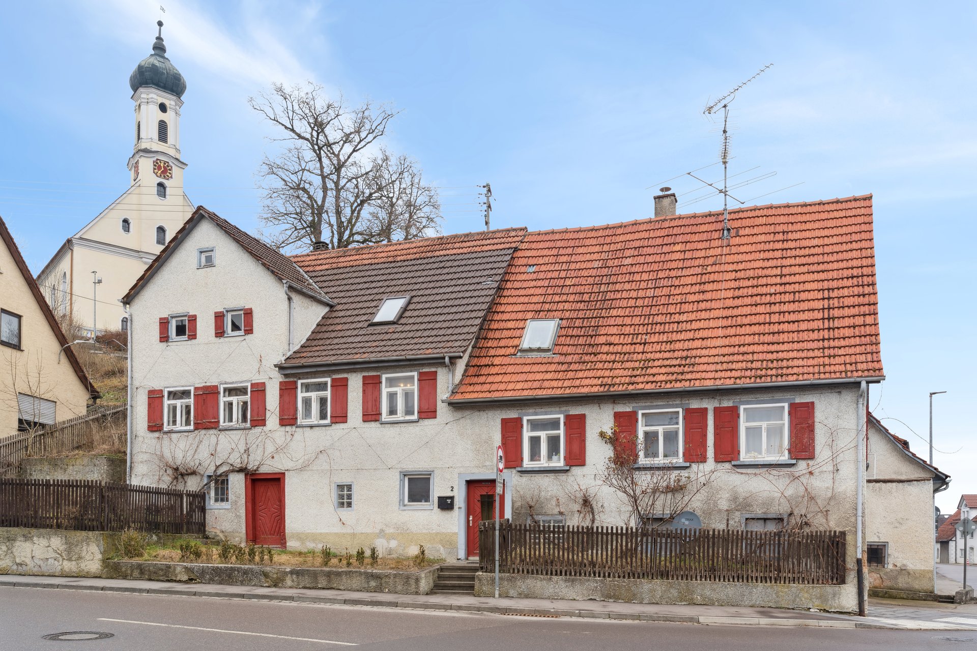 A two-story white house with red shutters and a tiled roof, situated next to a church with a tall steeple. [AI-generated text]