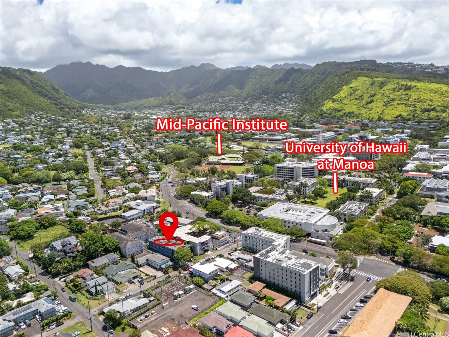 Aerial view of a neighborhood with houses, trees, and mountains in the background. A red pin marks a building near the University of Hawaii at Manoa.