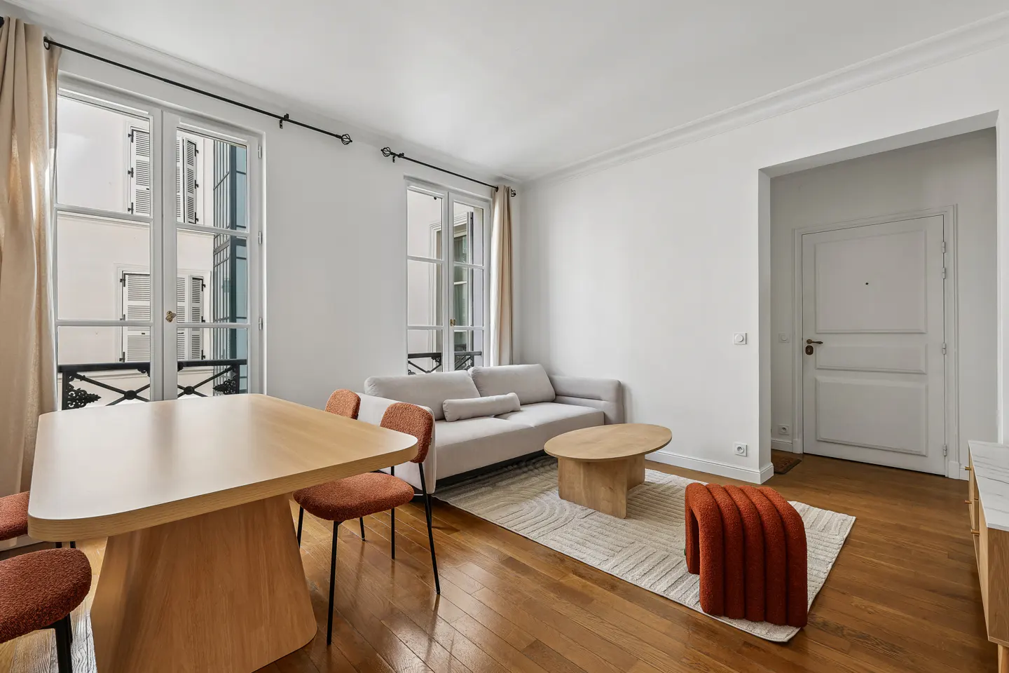 Bright living room with wood floors, white walls, and large windows. A light gray sofa, wood table, and orange chairs furnish the space.