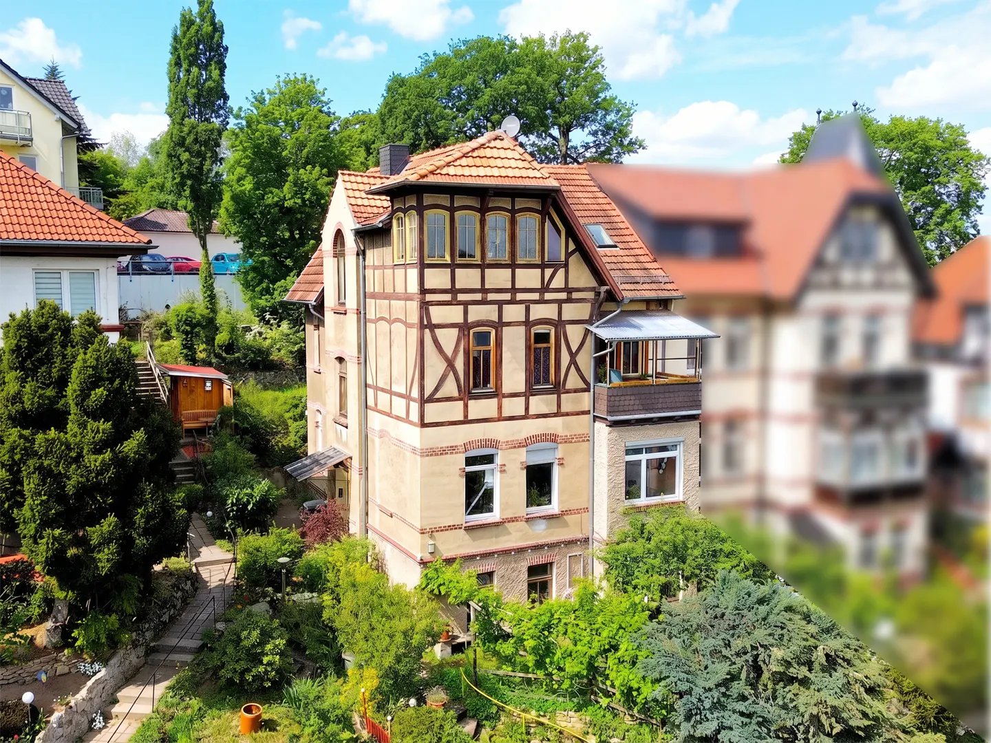 Exterior view of a three-story house with a red tile roof and half-timbered facade, surrounded by lush greenery.