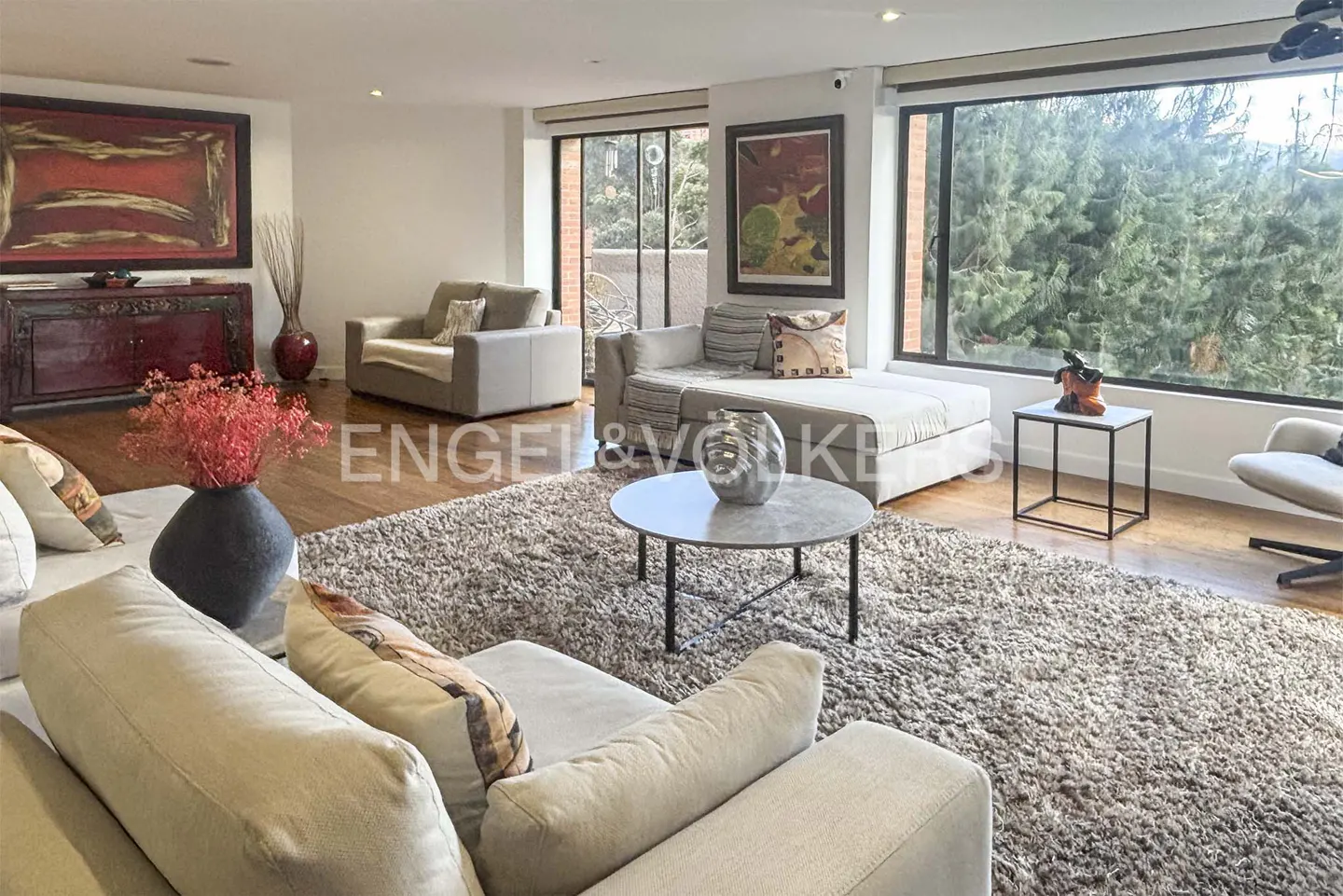A bright living room with beige sofas, a round table, and a large window overlooking trees. Artwork adorns the walls.