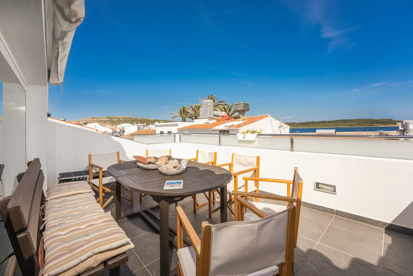 Sunny rooftop terrace with a dining table, chairs, and a bench, overlooking a coastal town under a clear blue sky.