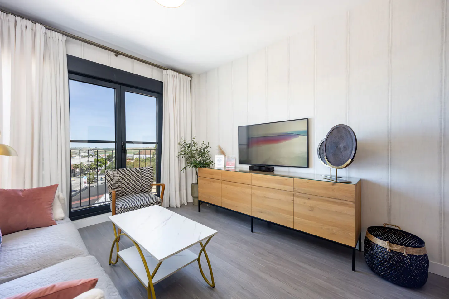 Bright living room with a white sofa, pink pillows, a marble coffee table, and a wooden TV stand with a flat-screen TV. A large window shows a city view.