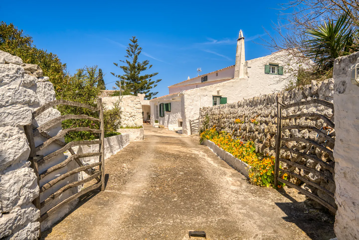 Gated driveway leading to a white house with green shutters under a blue sky.