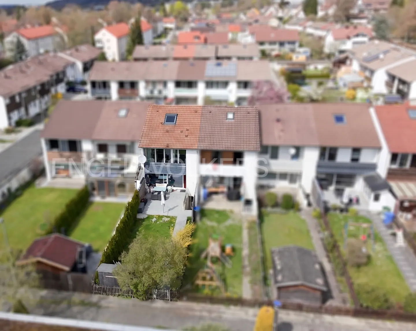 Aerial view of a row of townhouses with red tile roofs, green lawns, and a blurred background of trees and other houses.