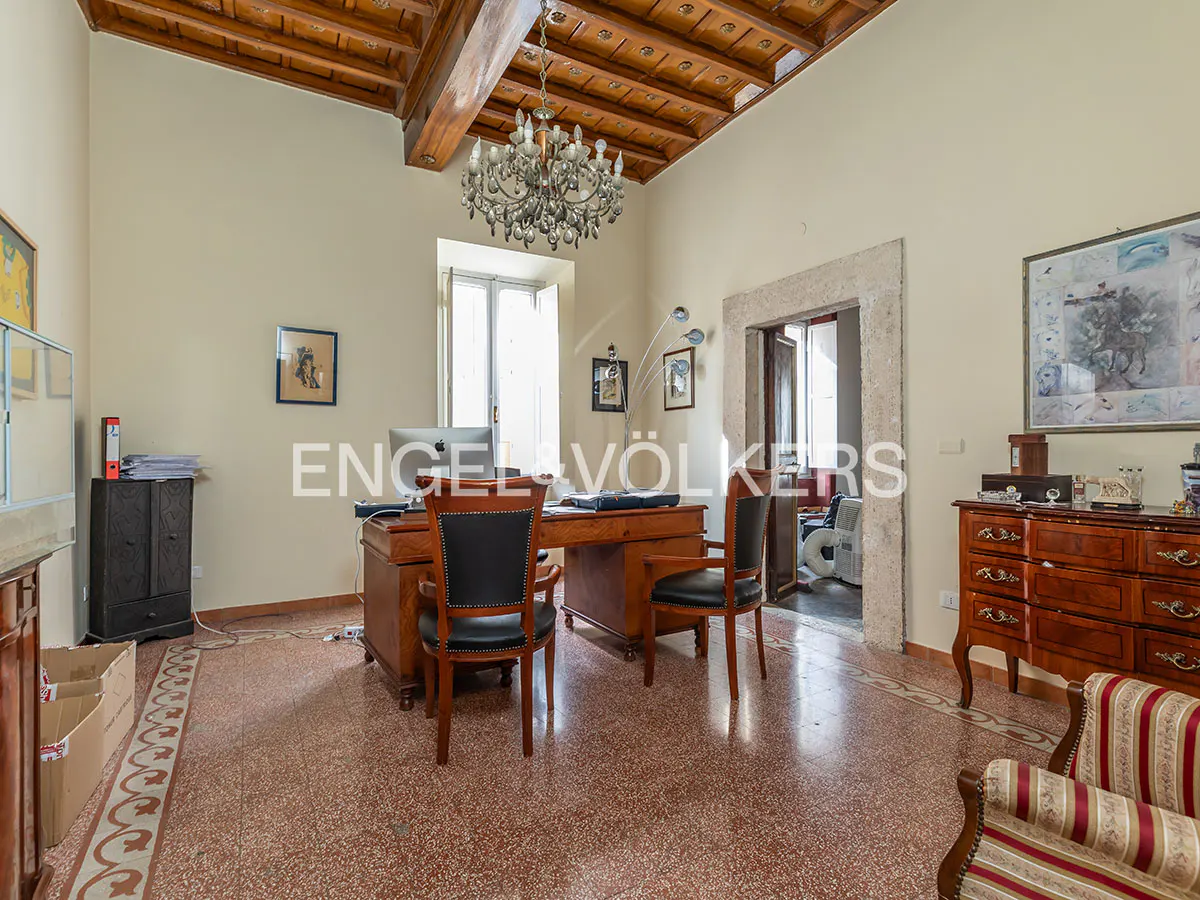 A spacious office with a wooden desk, two black chairs, and a chandelier. The room has a wooden ceiling and a patterned tile floor.